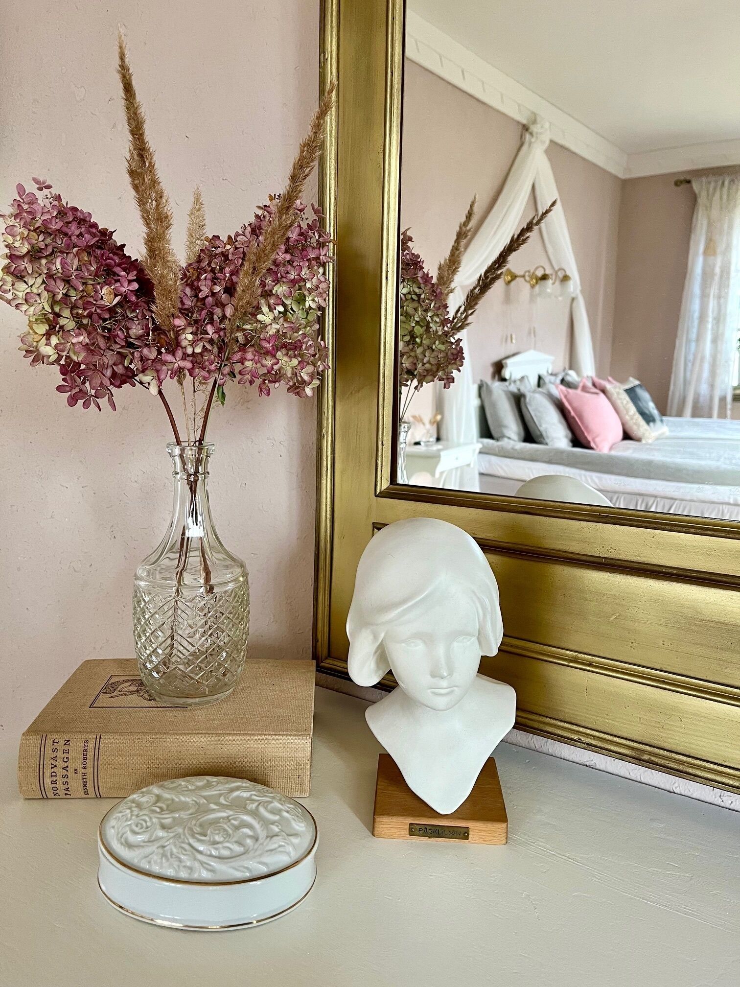 A room with a bed, a mirror, a vase with dried flowers, a bust, and a small decorative plate.