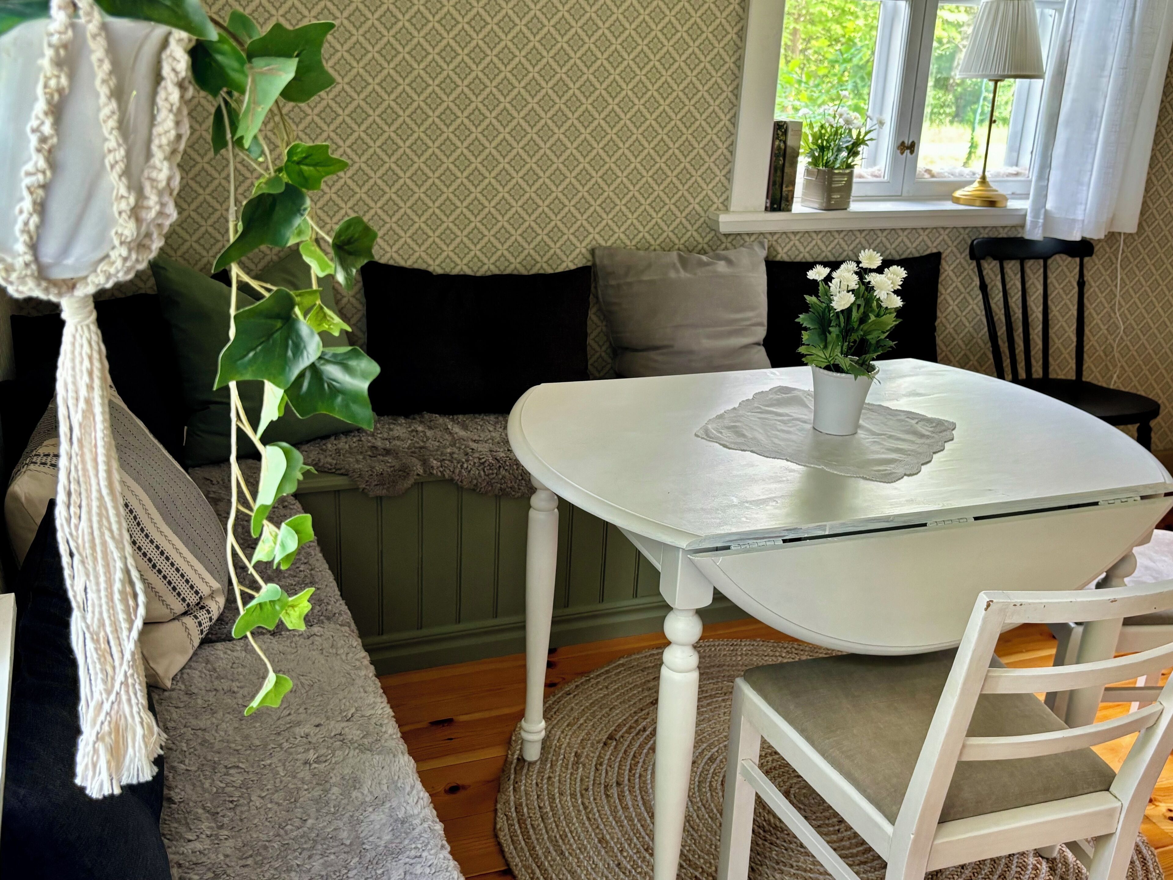 A cozy room with a white table, a cushioned bench, and a chair. There is a window with a view of greenery outside.