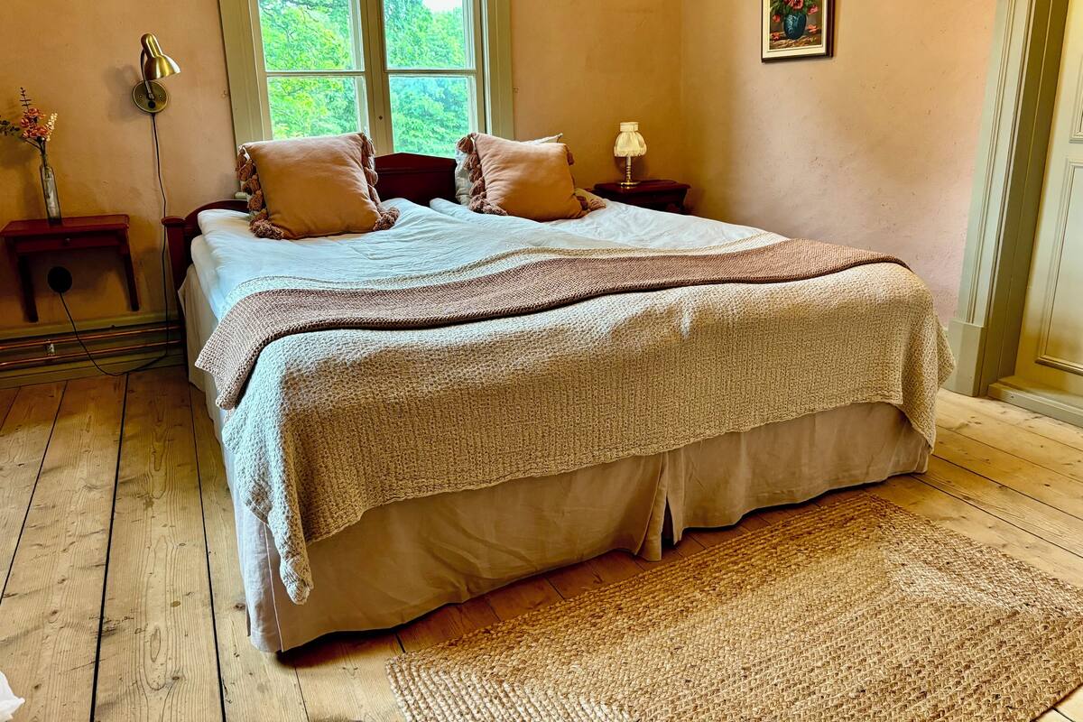 A bedroom with a large bed, a window with a view of greenery, a bedside table with a lamp, and a painting on the wall.