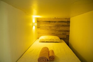 A small, single-bed room with a wooden headboard, a bedside lamp, and two rolled towels on the bed.