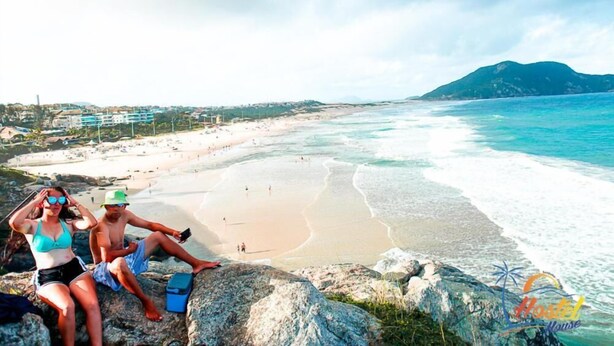 A beach with people sitting on rocks, a clear view of the ocean, and a distant mountain. - Hostel House Santinho (Florianópolis)