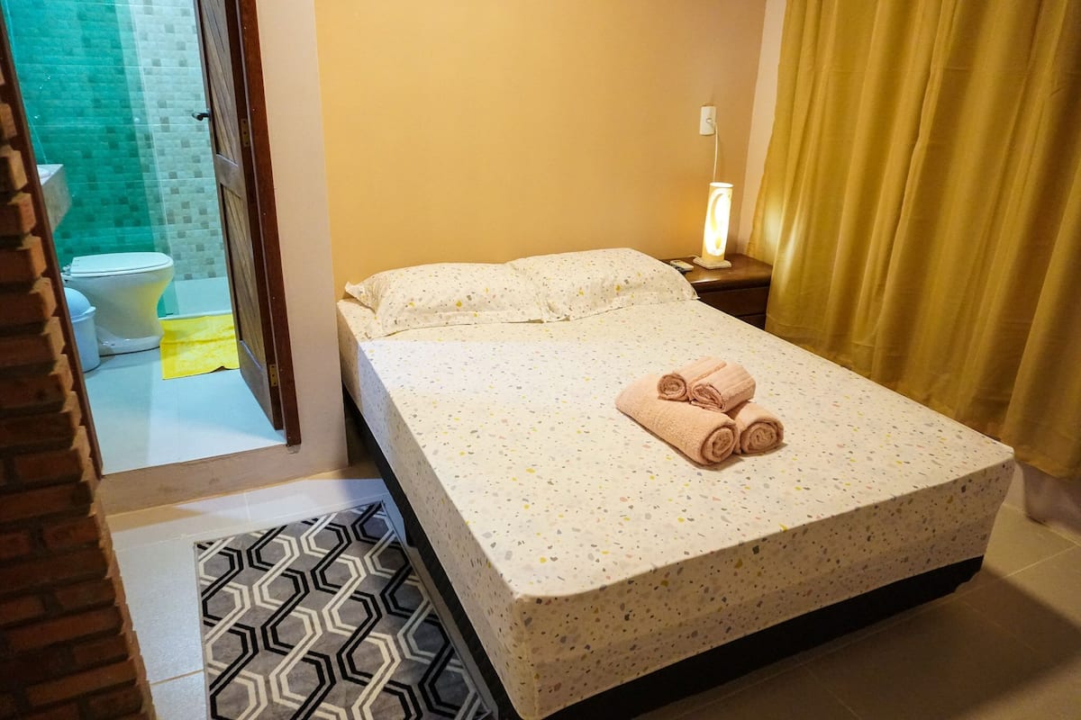 Ein Hotelzimmer mit Bett, Nachttisch, Lampe, und einem durch eine offene Tür einsehbaren Badezimmer sowie einem gemusterten Teppich.