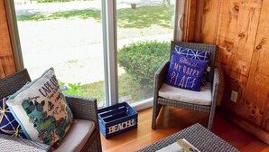 Een gezellige kamer met rieten meubels, een groot raam met uitzicht op een tuin, en een blauwe kist met de tekst 'BEACH'.