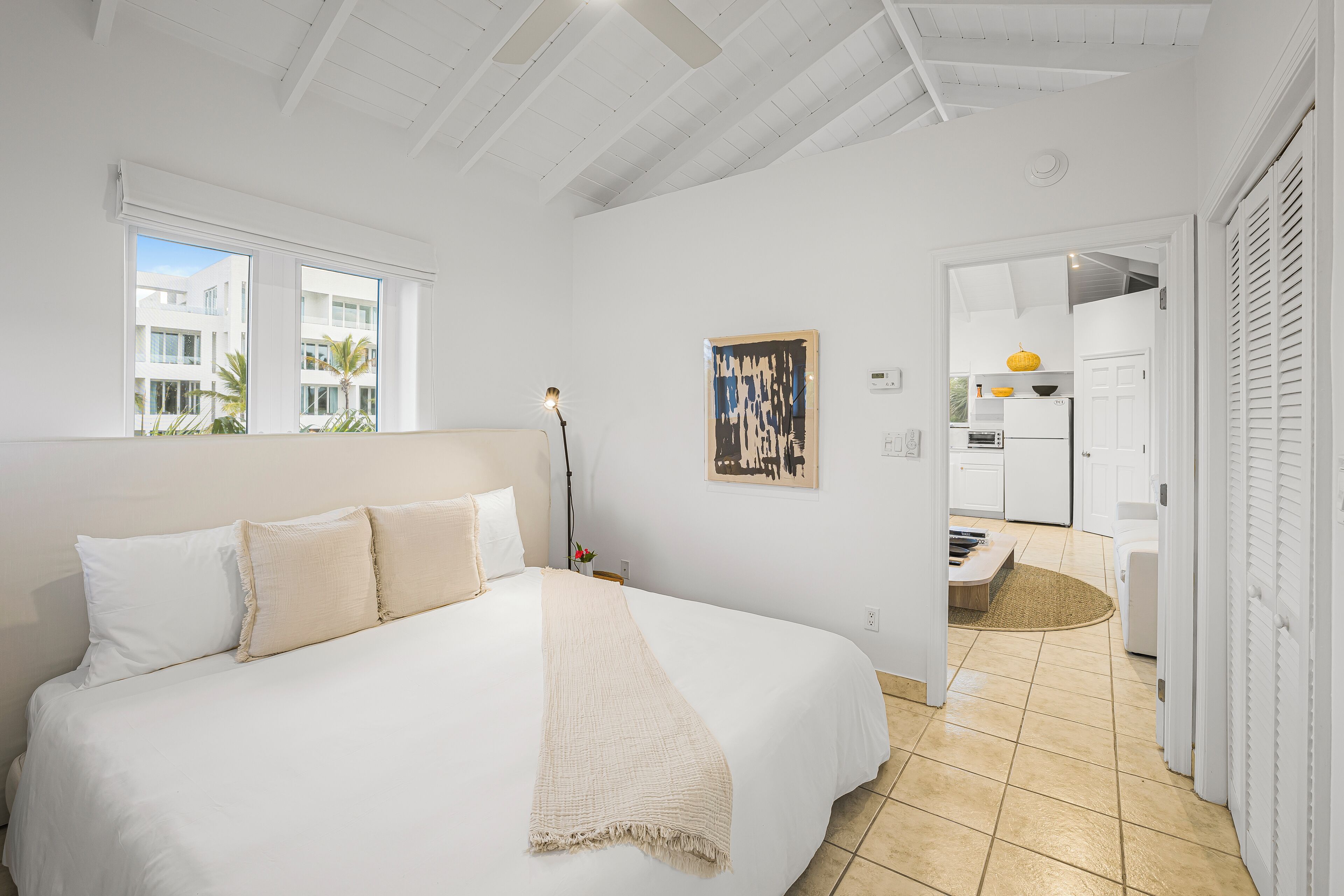 A bedroom with a large bed, a window, a painting, and a view of the outside.