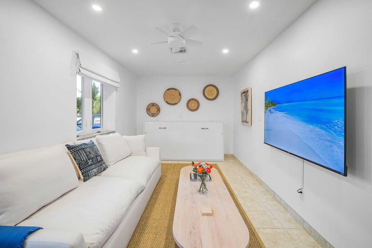 Un salon moderne avec un canapé blanc, une table basse ornée d’un vase de fleurs, un téléviseur à écran plat fixé au mur et un ventilateur de plafond.