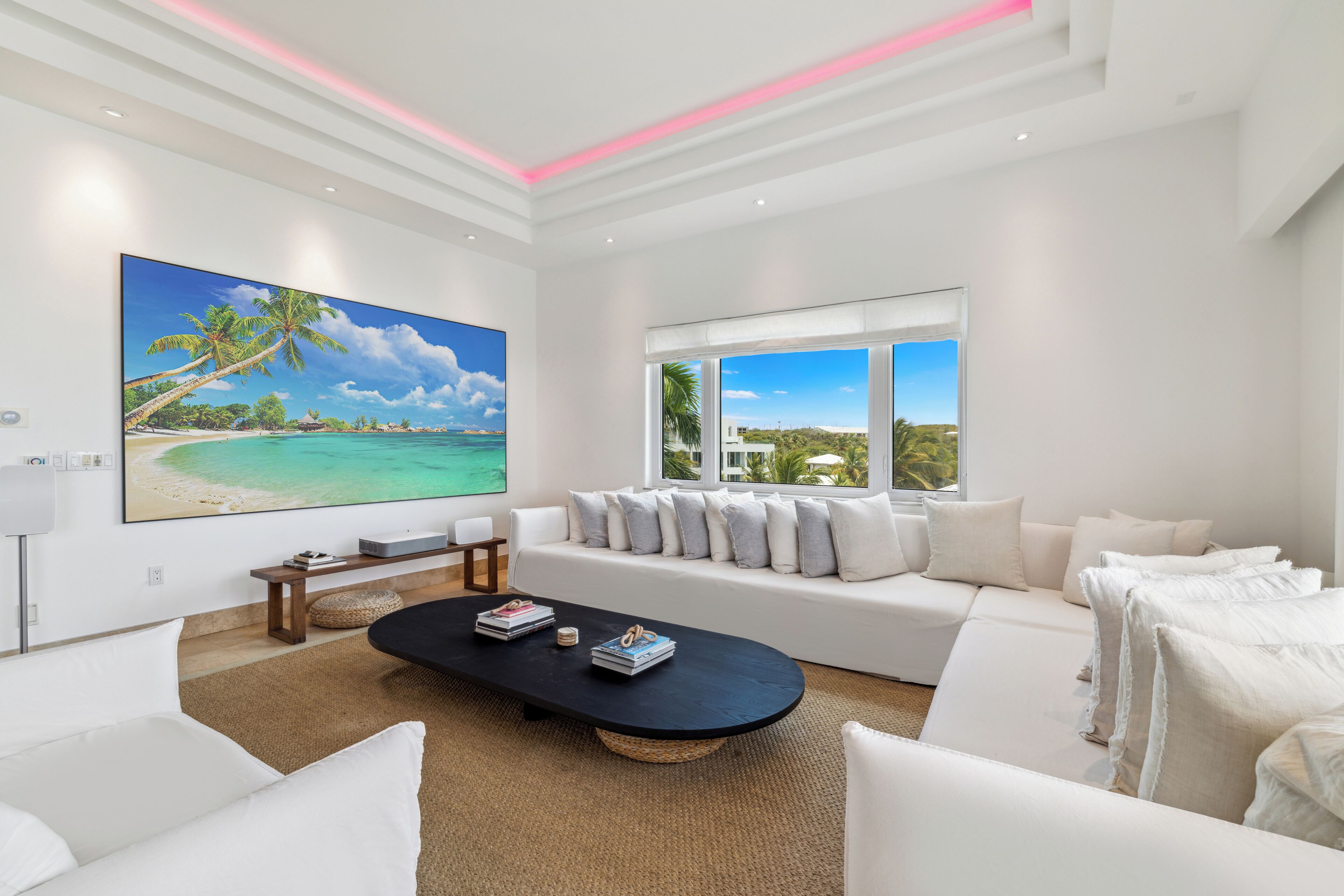 A modern living room with a large flat-screen TV, a black coffee table, white sofas, and a large window with a view of the beach.