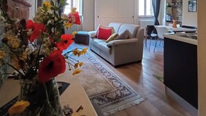Een ruime woonkamer met een bank, een salontafel en een vaas met bloemen. Er is een keukenhoek met een aanrecht en een spoelbak.