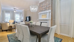 A dining area with a dark wooden table, white upholstered chairs, a chandelier, a brick accent wall, and a fireplace.