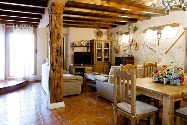 A rustic living room with a wooden dining table, two sofas, a TV, and a stone wall with mounted tools.