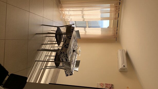 Een eethoek met een tafel en stoelen, een raam met gordijnen, en een wandairconditioner.