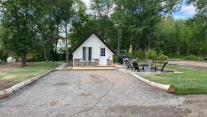 Eine kleine weiße Holzhütte mit Steinfundament, einer Veranda mit zwei Stühlen und einem überdachten Bereich mit Tisch und Stühlen.