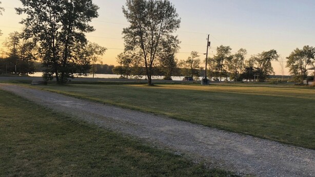 A grassy area with a gravel path, trees, and a body of water in the background. - Cozy little cottage with big memories (Campbellford)