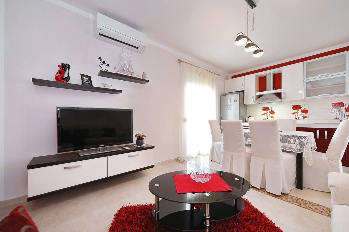 Een moderne woonkamer met een flatscreen televisie, een eetgedeelte met een ronde tafel en een keuken met witte kasten en rode accenten.