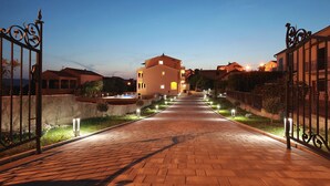 Un chemin pavé mène à un bâtiment doté d’une piscine, qui est entouré de clôtures bien éclairées et de verdure.