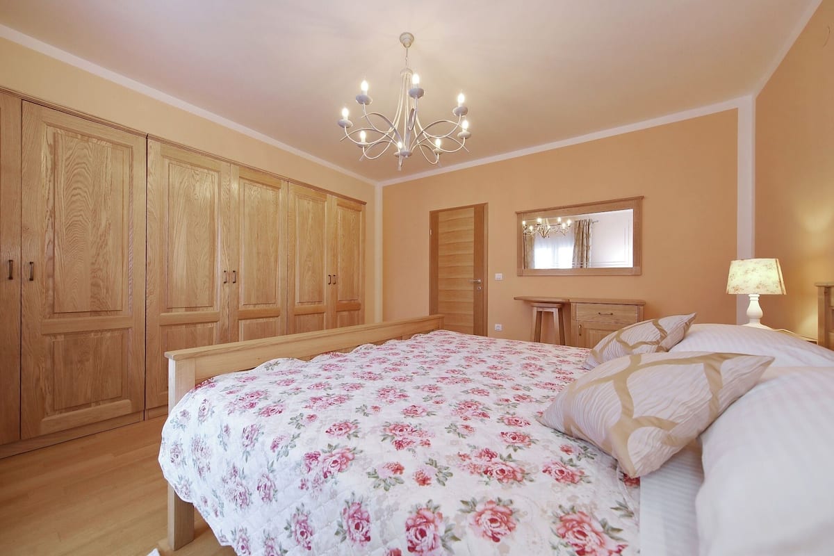 Een slaapkamer met een groot bed, een houten garderobe, een kroonluchter, een spiegel en een nachtkastje met een lamp.