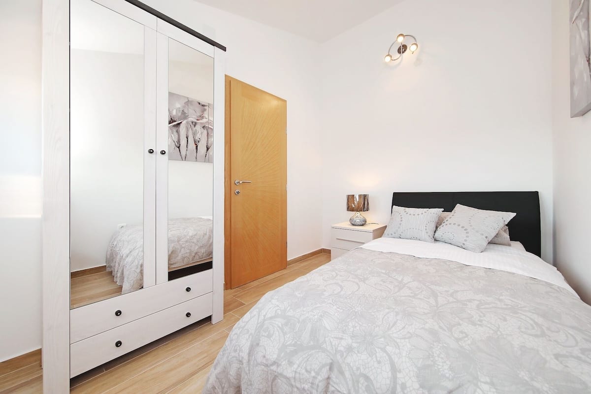 Een slaapkamer met een bed, een kledingkast met spiegel, een nachtkastje met lamp en een deur.