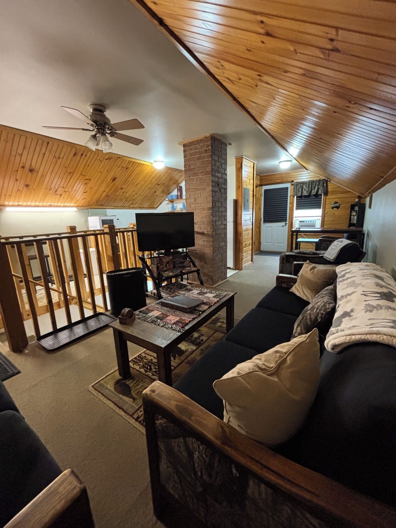 A cozy living room with a wooden ceiling, a television on a stand, a sofa with cushions, and a small table with a rug.