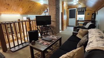 A cozy living room with a wooden ceiling, a television on a stand, a sofa with cushions, and a small table with a rug.
