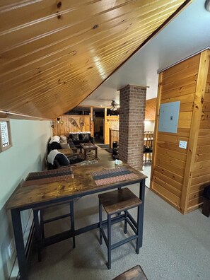A cozy room with a wooden ceiling, a brick fireplace, a dining table with chairs, and a seating area with couches.