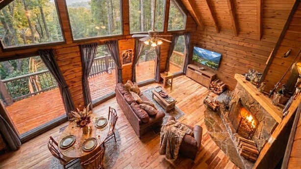 Interior de una cabaña de madera con una amplia mesa de comedor, sofá, chimenea y grandes ventanales con vistas al bosque.