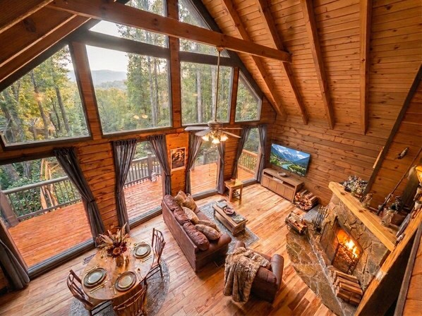 A wooden cabin interior with a large dining table, sofa, fireplace, and large windows offering a forest view.