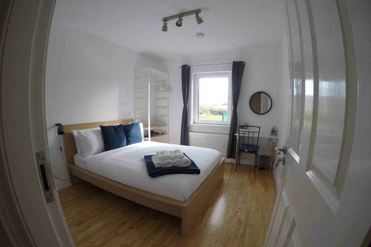 Quarto com cama de madeira, mesa de cabeceira, janela com cortinas, radiador e espelho na parede.