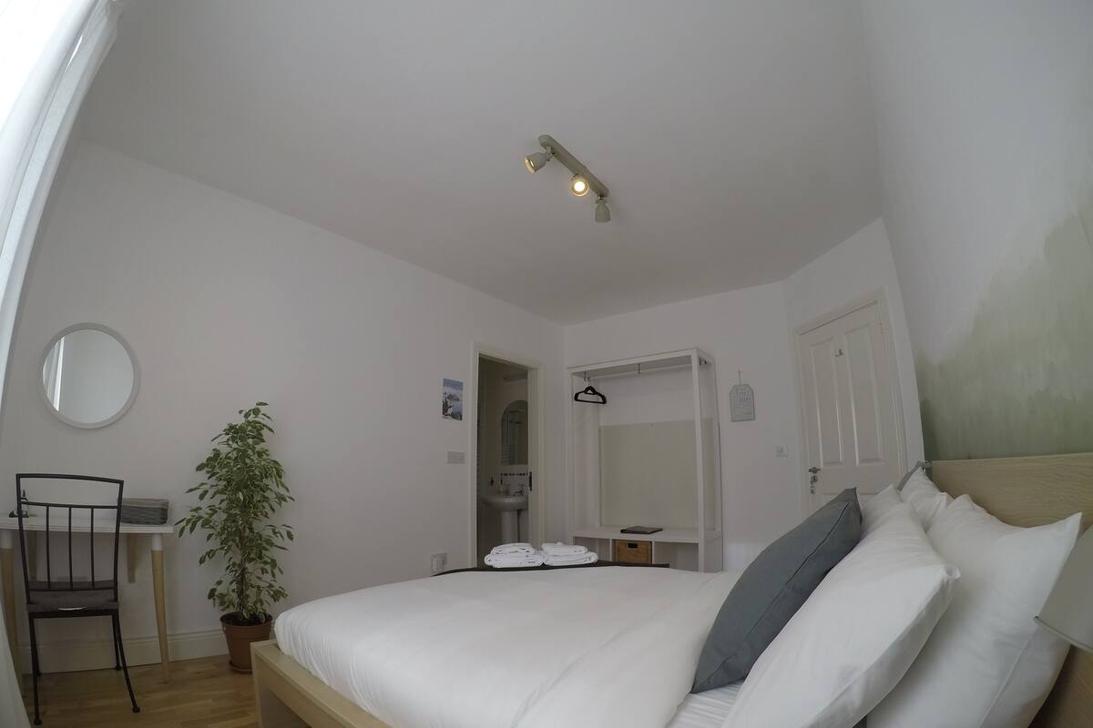 Quarto com cama, uma cadeira, planta em vaso, escrivaninha e luminária pendente.