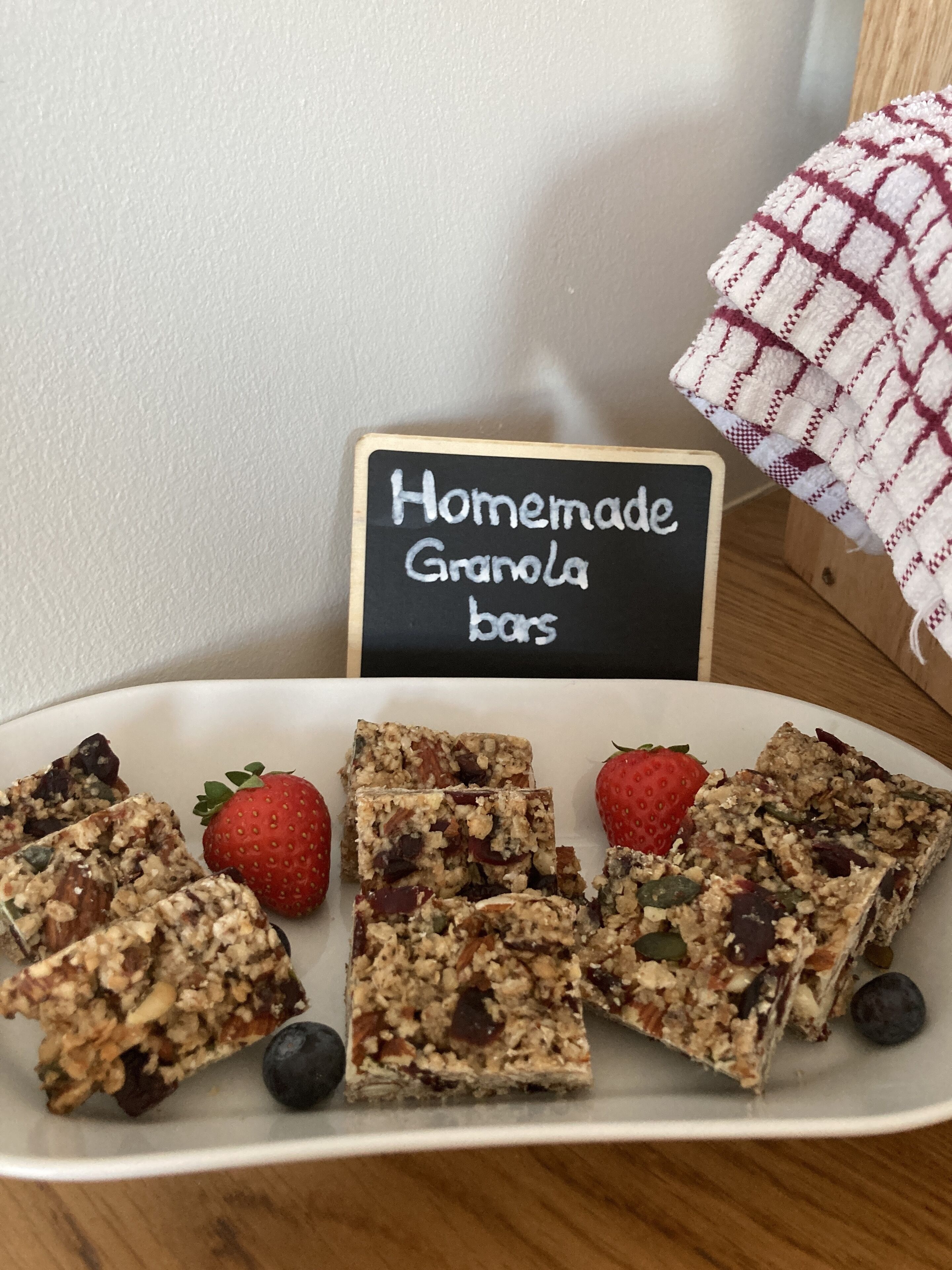Ein Teller mit hausgemachten Granola-Riegeln, Erdbeeren und Blaubeeren, ein Kreidetafel mit der Aufschrift 'Handgemachte Granola-Riegel' und ein kariertes Tuch im Hintergrund.