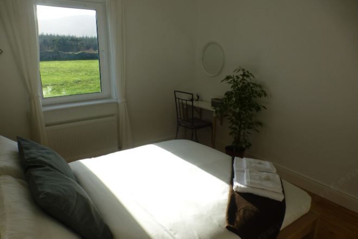 Un dormitorio con cama, ventana con vistas a un campo verde, escritorio con silla y una planta en maceta.