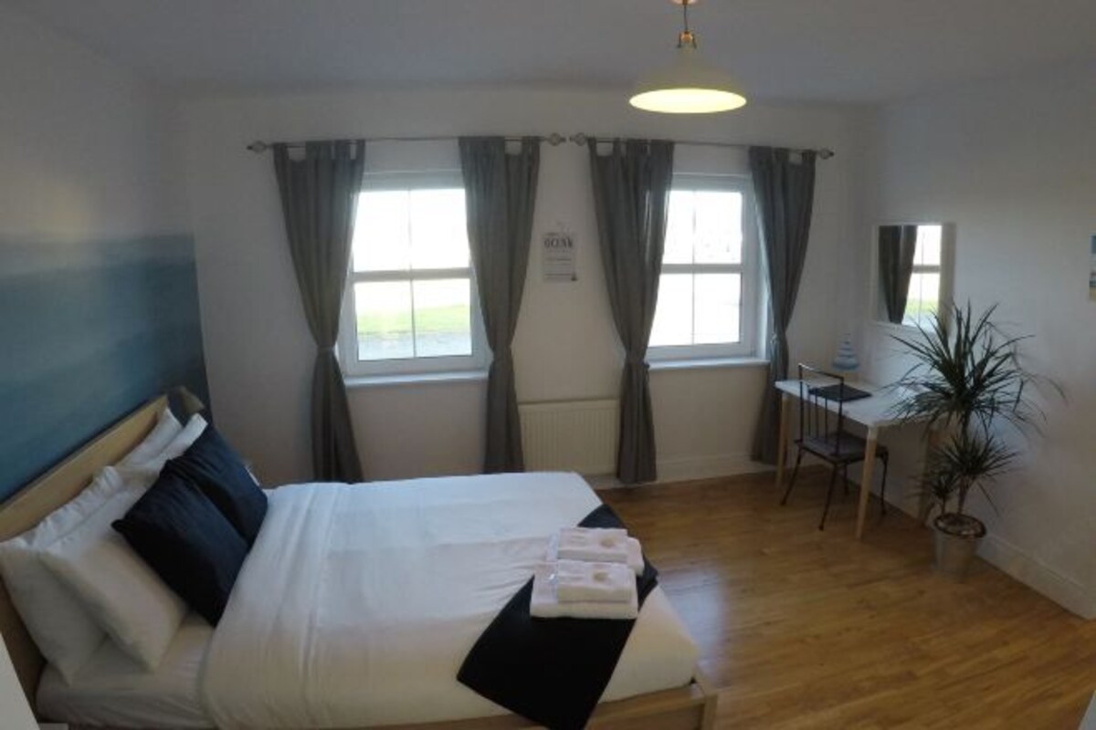 Habitación de hotel con una cama, un escritorio y una silla. Hay dos ventanas con cortinas, una planta en maceta y un cartel en la pared.