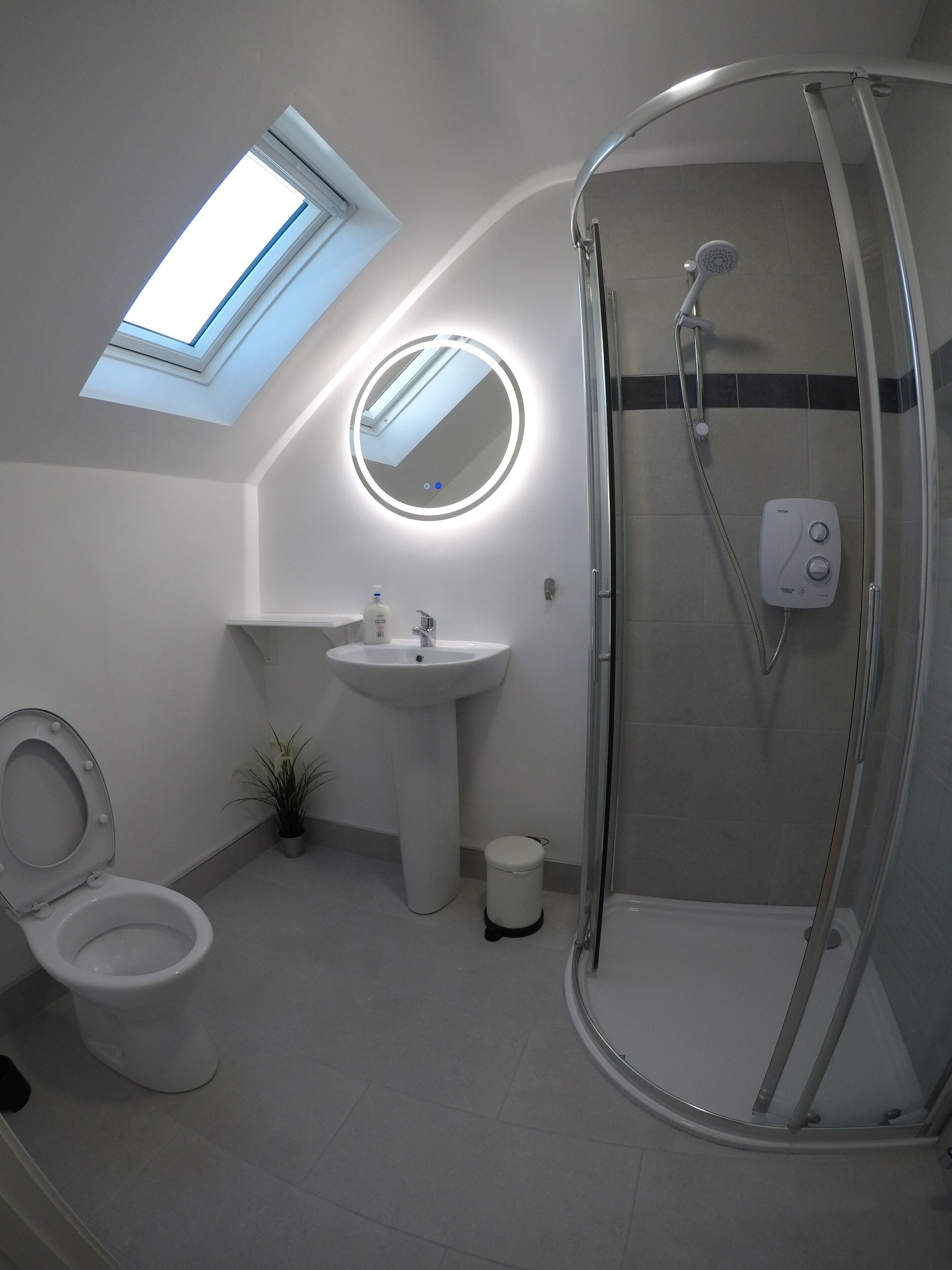 Et moderne badeværelse med ovenlys, et rundt spejl, en vask, et toilet og en brusekabine med glasvægge.