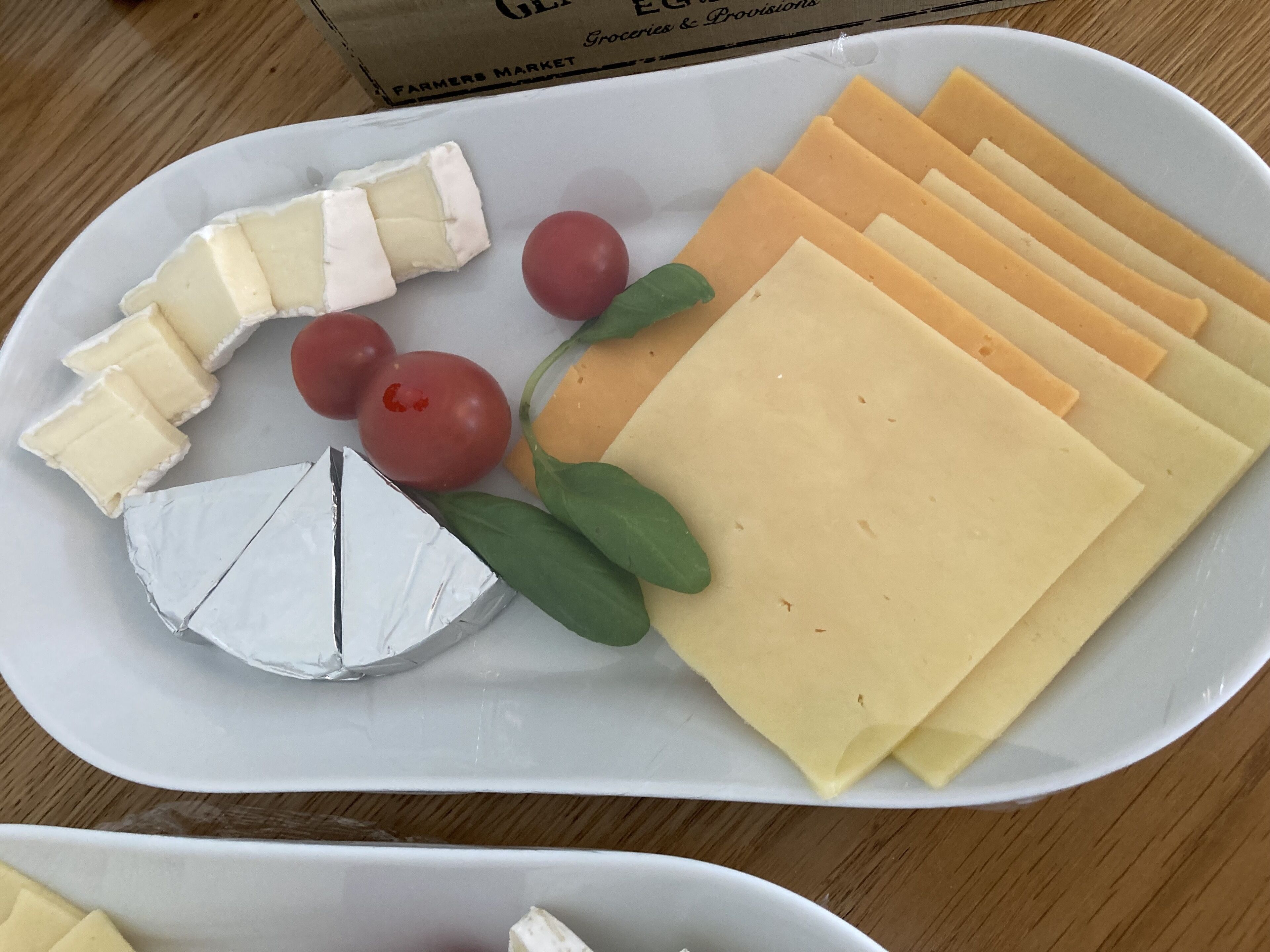 Une assiette garnie d’une variété de fromages, dont un morceau de brie et plusieurs tranches de cheddar, accompagnée de tomates cerises et d’une branche de verdure.