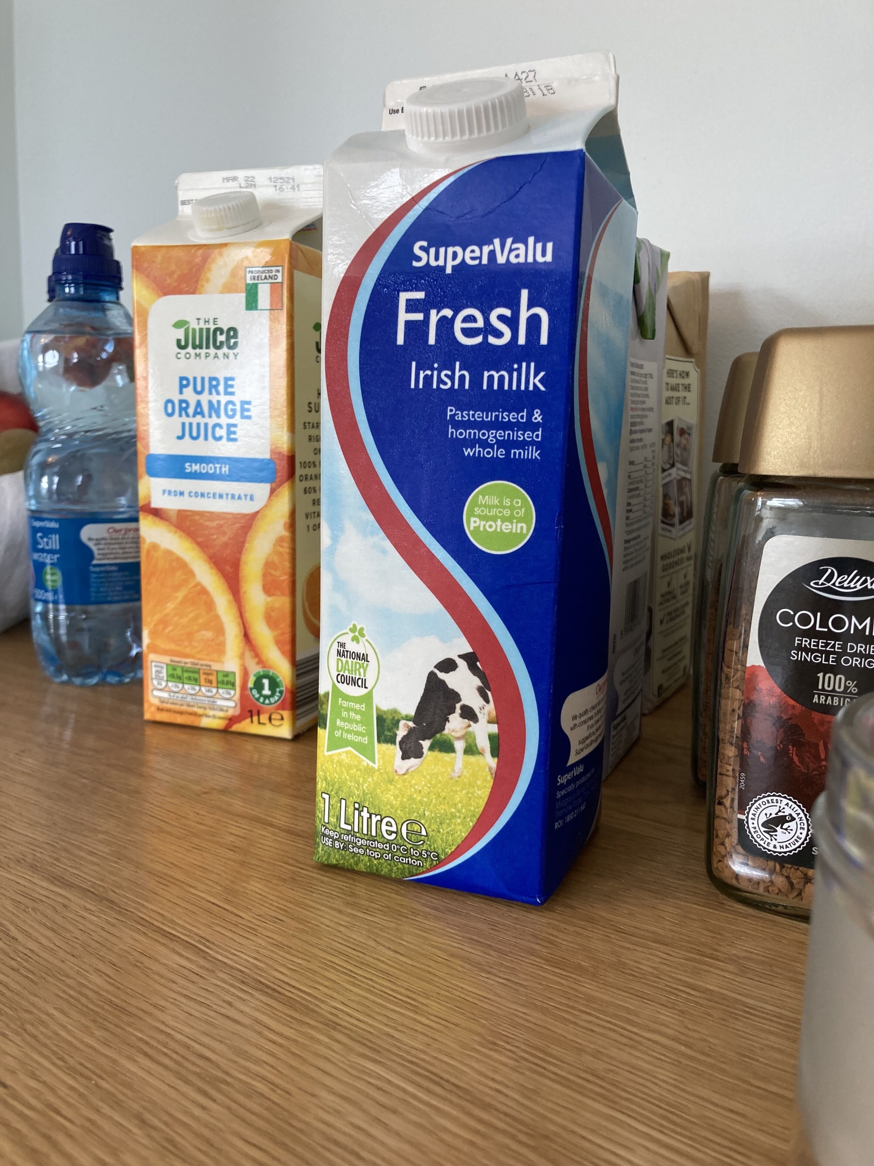 Een karton SuperValu Fresh Ierse melk, een fles Pure sinaasappelsap en een fles stilstaand water op een tafel.