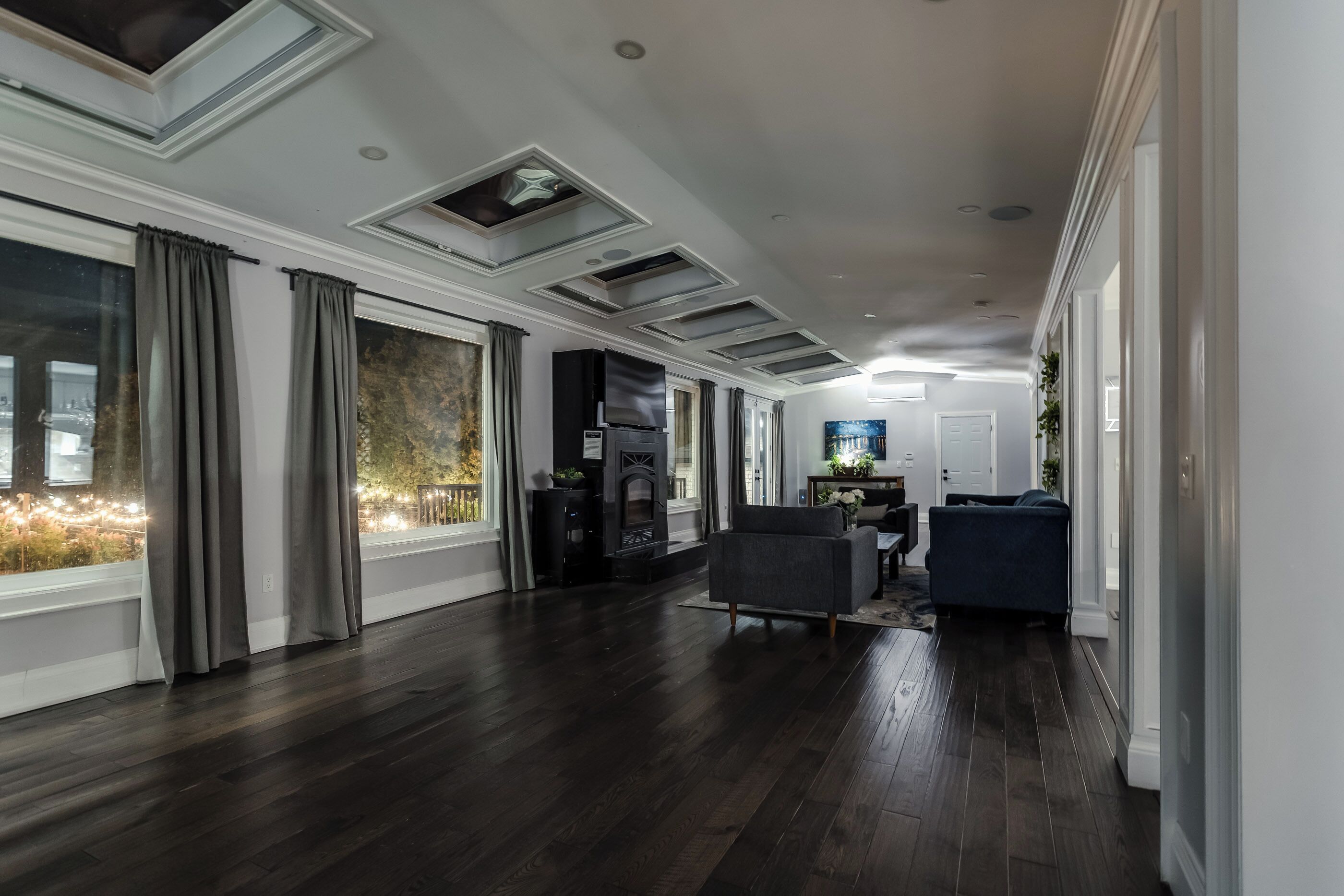 Amplia sala de estar con piso de madera oscura, ventanas grandes con cortinas, chimenea y zona de estar con sofá y sillones.