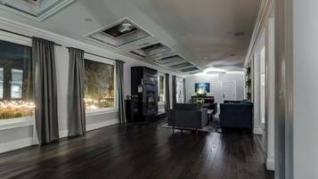 Amplia sala de estar con piso de madera oscura, ventanas grandes con cortinas, chimenea y zona de estar con sofá y sillones.