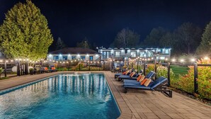 Une piscine entourée d’une terrasse en bois et d’un bâtiment doté de nombreuses fenêtres, avec des chaises longues et des guirlandes lumineuses.