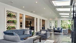Un salon spacieux avec un grand canapé d’angle gris, une table basse et une cheminée. On y trouve plusieurs options d’assises, notamment des fauteuils et un canapé. La pièce est dotée d’une grande fenêtre offrant une vue sur la verdure environnante.