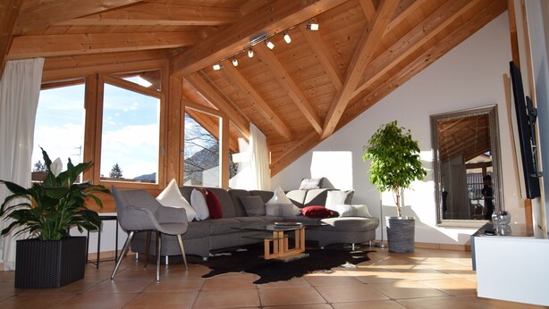 A spacious living room with a high ceiling, large windows, a sofa, chairs, a coffee table, and a potted plant. - Favorite place Apartment 2 Arrive and feel good (Garmisch-Partenkirchen)