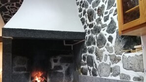 A stone fireplace with a fire, a wooden cabinet, and a cushioned chair in a room with a stone wall. - Vacation Home Pico Açôres (Calheta de Nesquim)