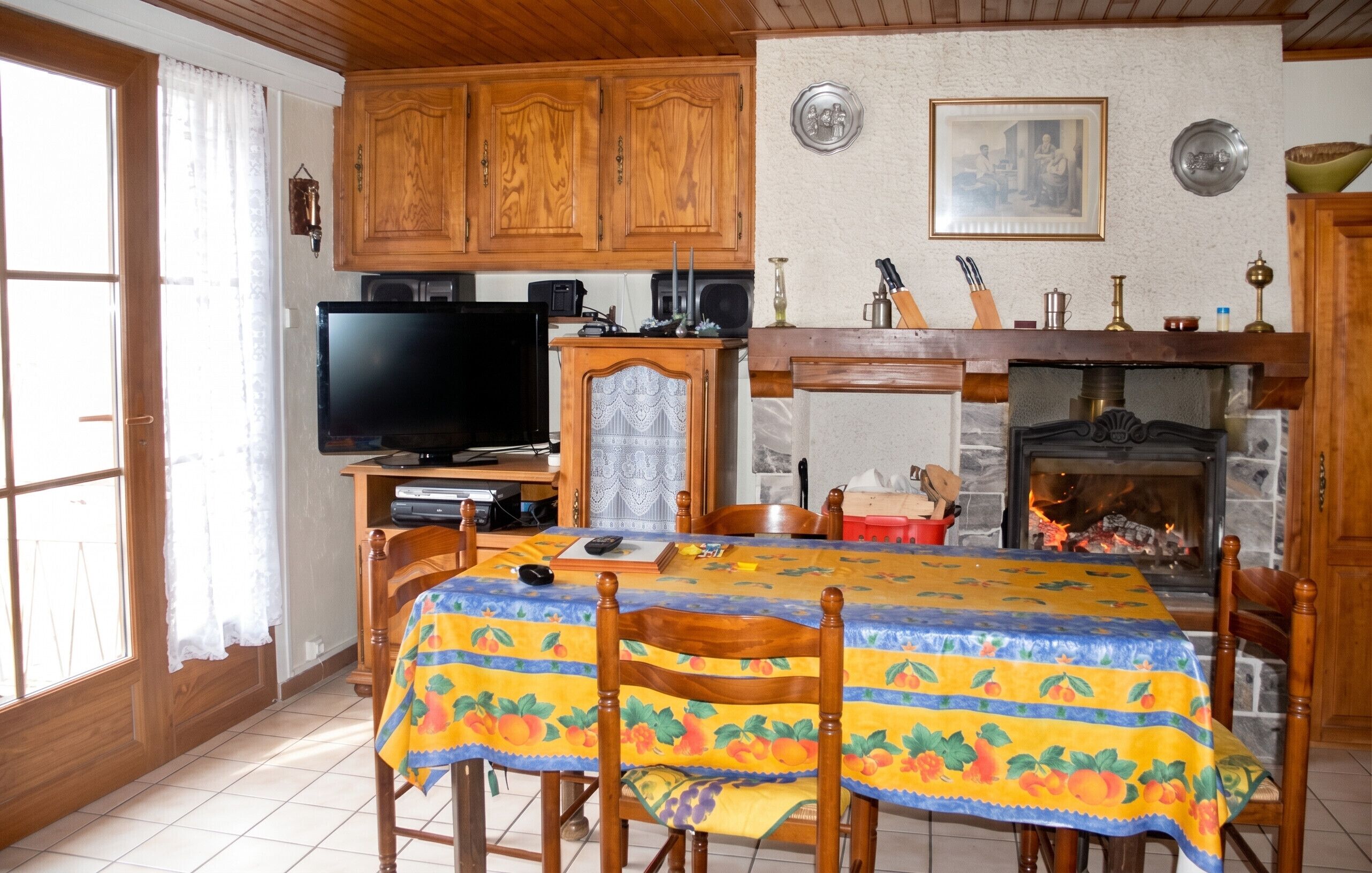 Uma área de refeições com uma mesa coberta por um guardanapo estampado, cadeiras de madeira, uma televisão, lareira e armários de madeira.