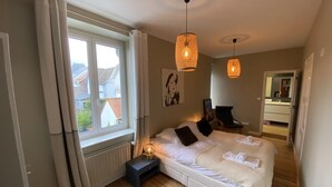 Ein Schlafzimmer mit Bett, Nachttisch, zwei Pendelleuchten, einem Sessel, einem Fenster mit Vorhängen und Blick nach draußen.