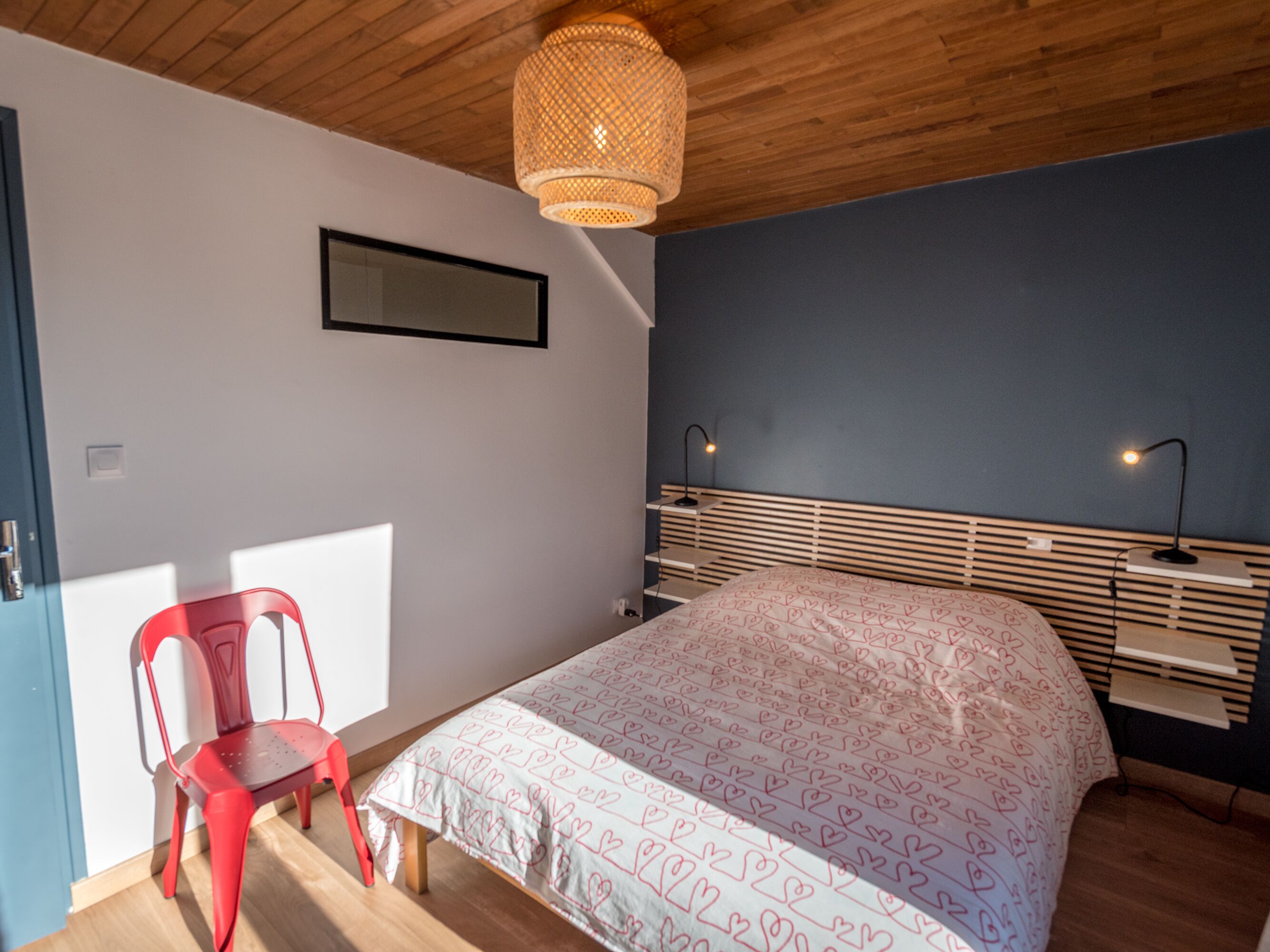 A bedroom with a bed, a red chair, a bedside table, a lamp, and a hanging light fixture.