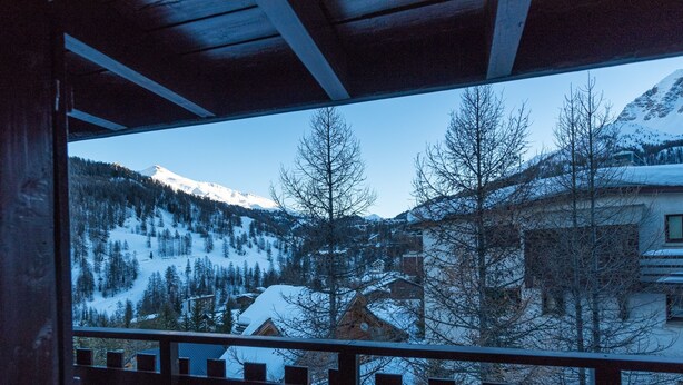 A view from a balcony with a wooden railing, overlooking a snow-covered mountain landscape with buildings and trees.