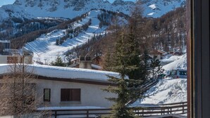 A mountain landscape with snow-covered peaks, a ski slope, and a building with a wooden deck in the foreground.