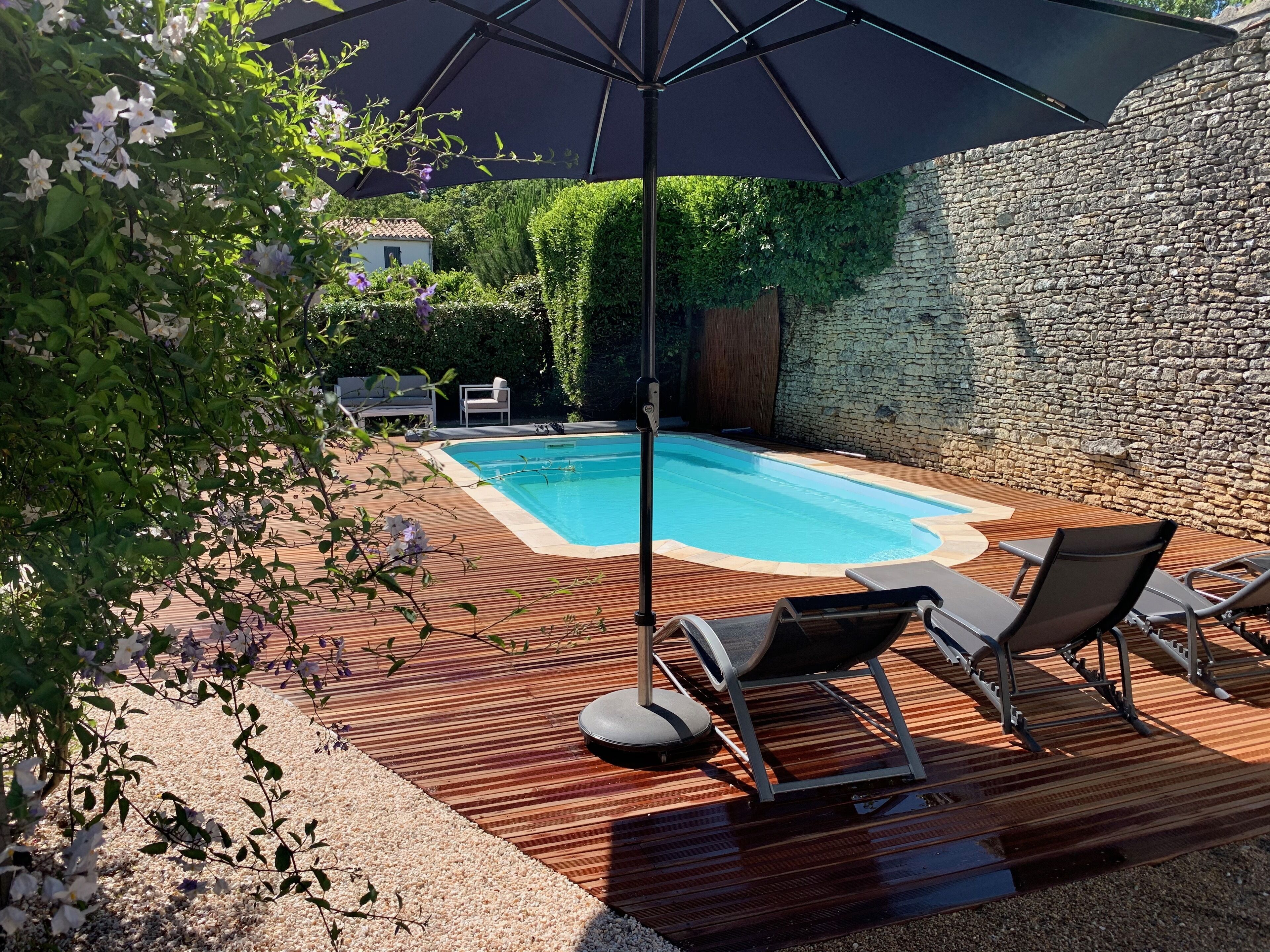 A swimming pool with wooden decking and lounge chairs, surrounded by greenery and a stone wall.