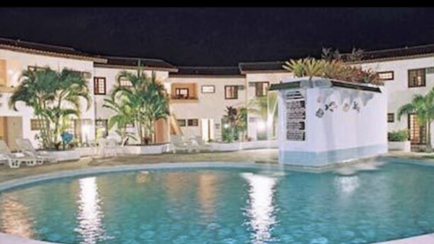 A swimming pool surrounded by a building with multiple balconies, palm trees, and a well-lit pool area. - Apartamento - La Corte (Porto Seguro)