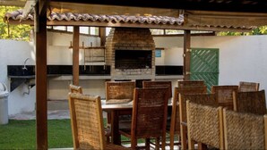 An outdoor patio with a brick pizza oven, wooden chairs, and a table. - Apartamento - La Corte (Porto Seguro)