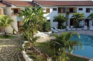 A poolside area with lounge chairs, palm trees, and a building with a tiled roof. - Apartamento - La Corte (Porto Seguro)