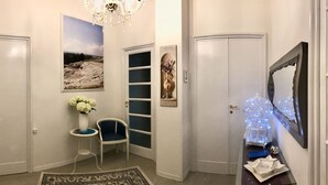 A hallway with white doors, a chandelier, a framed landscape photo, a small white chair, a vase with flowers, and a shelf with decorative items. - Alice's House Luxury Hospitality - Elevator & Fast Wi-fi (Siracusa)