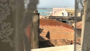 A view through a window with lace curtains, revealing a coastal town with terracotta rooftops and buildings with balconies. - Alice's House Luxury Hospitality - Elevator & Fast Wi-fi (Siracusa)